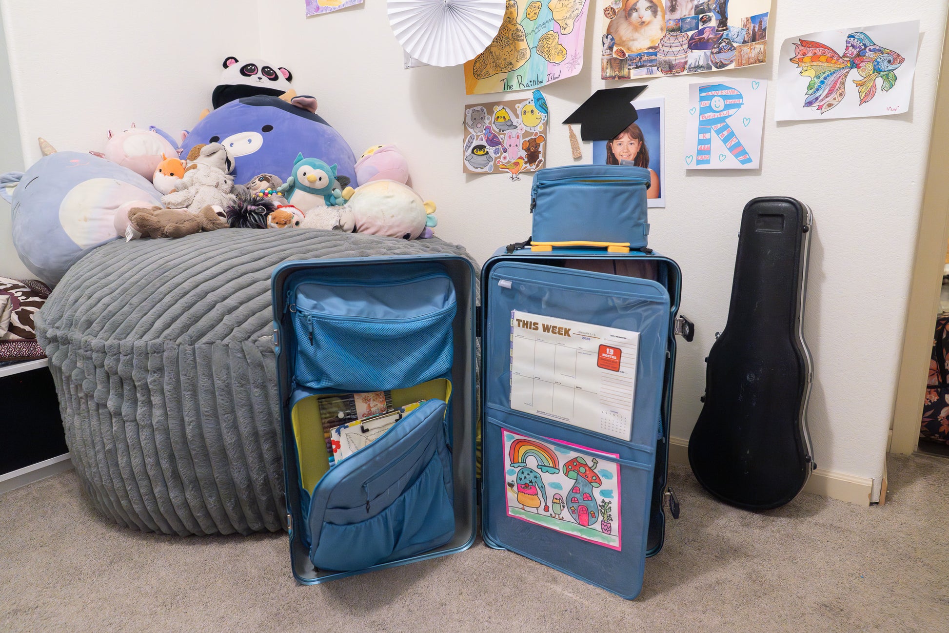 Open blue Belonging LifeLocker Founders Bundle displayed in a child’s bedroom beside a bed with stuffed animals, showing interior compartments with a calendar and artwork, and a removable organizer placed on top.