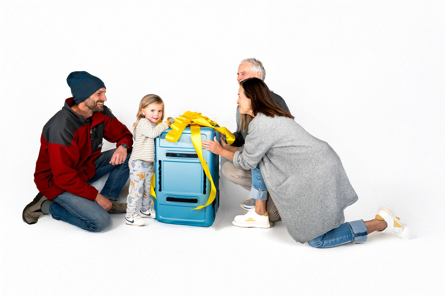 Family gathered around a blue Belonging LifeLocker Founders Bundle suitcase with a large yellow bow, smiling as a young child stands beside it on a white studio background.
